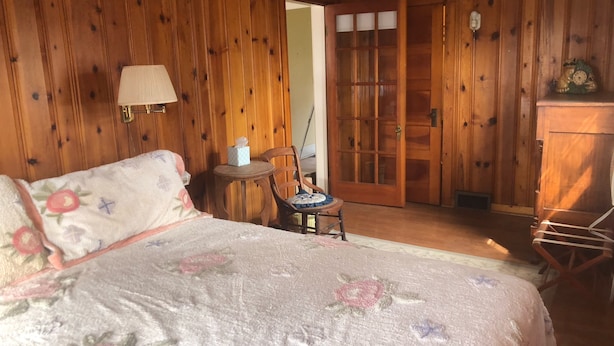 Una habitación con paneles de madera, una cama, una silla, una mesita y una puerta.