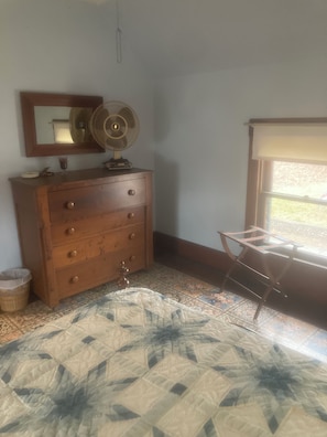 A room with a wooden dresser, a standing fan, a mirror, a window with a view, and a patterned carpet.
