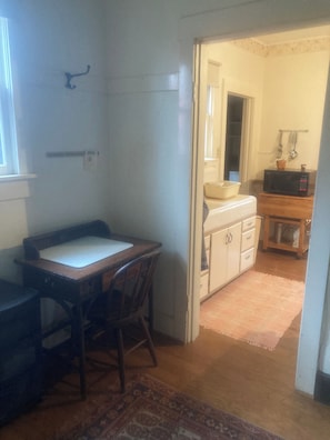 A room with a wooden desk, a chair, a small cabinet, and a microwave on a wooden shelf.