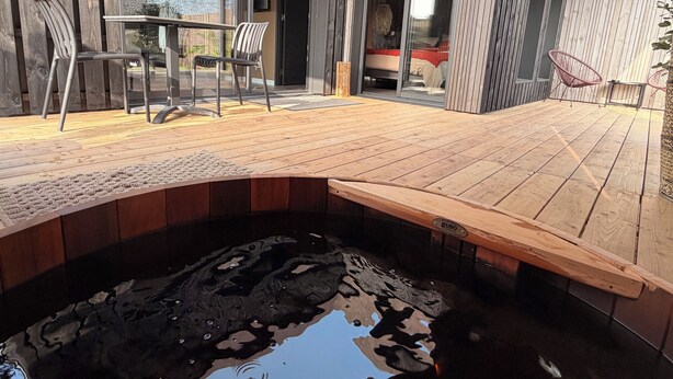 Une terrasse en bois avec un jacuzzi, des chaises et une porte vitrée donnant sur une pièce.