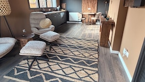 Une cuisine moderne avec des meubles de couleur sombre, un coin repas avec une table et des chaises en bois, et un espace salon confortable agrémenté d’un tapis à motifs.