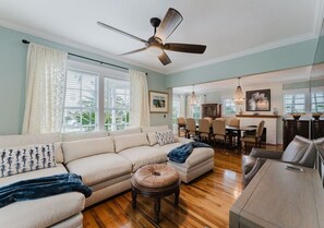 A living room with a sofa, ottoman, dining table, and ceiling fan. - Charleston House | Heated Pool and Screened Porch (West Palm Beach)