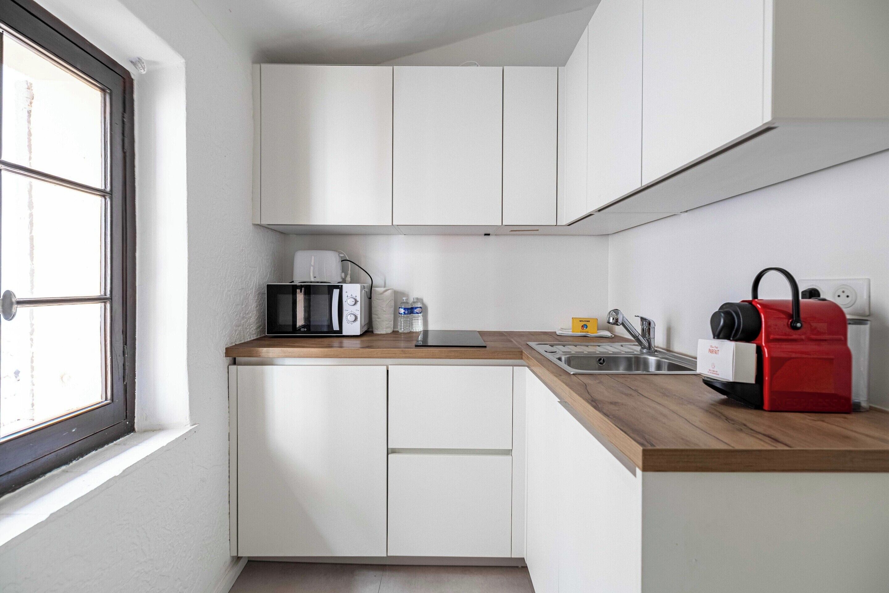 Una cocina compacta con armarios blancos, una encimera de madera, una cafetera, un hervidor y un electrodoméstico rojo, probablemente una aspiradora.