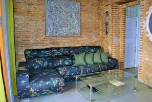 A living room with a brick wall, a camouflage sofa, a glass coffee table, and a decorative metal piece. - El Encanto de La Palma (Cádiz)