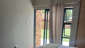 A bedroom with a large bed, a window with curtains, a brick wall, and a power outlet. - Room-serene Guest Room Retreat in Albert (Alberton)