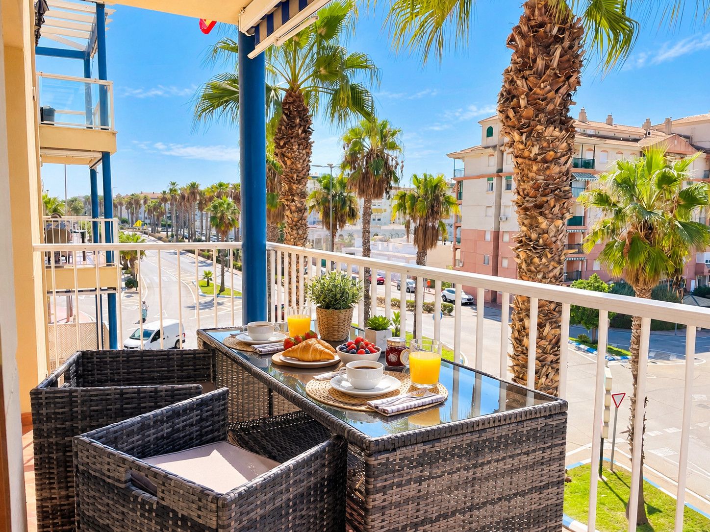 A balcony with wicker furniture, a table with breakfast items, and a view of palm trees and buildings.