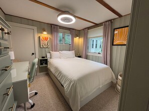 Ein Schlafzimmer mit Bett, Schminktisch, Nachttisch, Lampe und einem Fenster mit Vorhängen.
