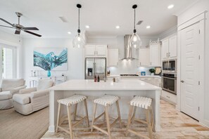A modern kitchen with white cabinets, a large island with stools, a ceiling fan, pendant lights, and a 'beach' sign on the wall.