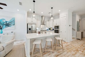 A modern kitchen with white cabinetry, a large island with stools, pendant lighting, and a stainless steel range hood.