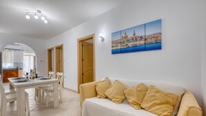En stue med en sofa, et spisebord med stole, et køkken i baggrunden og et bybillede som billedkunst på væggen.