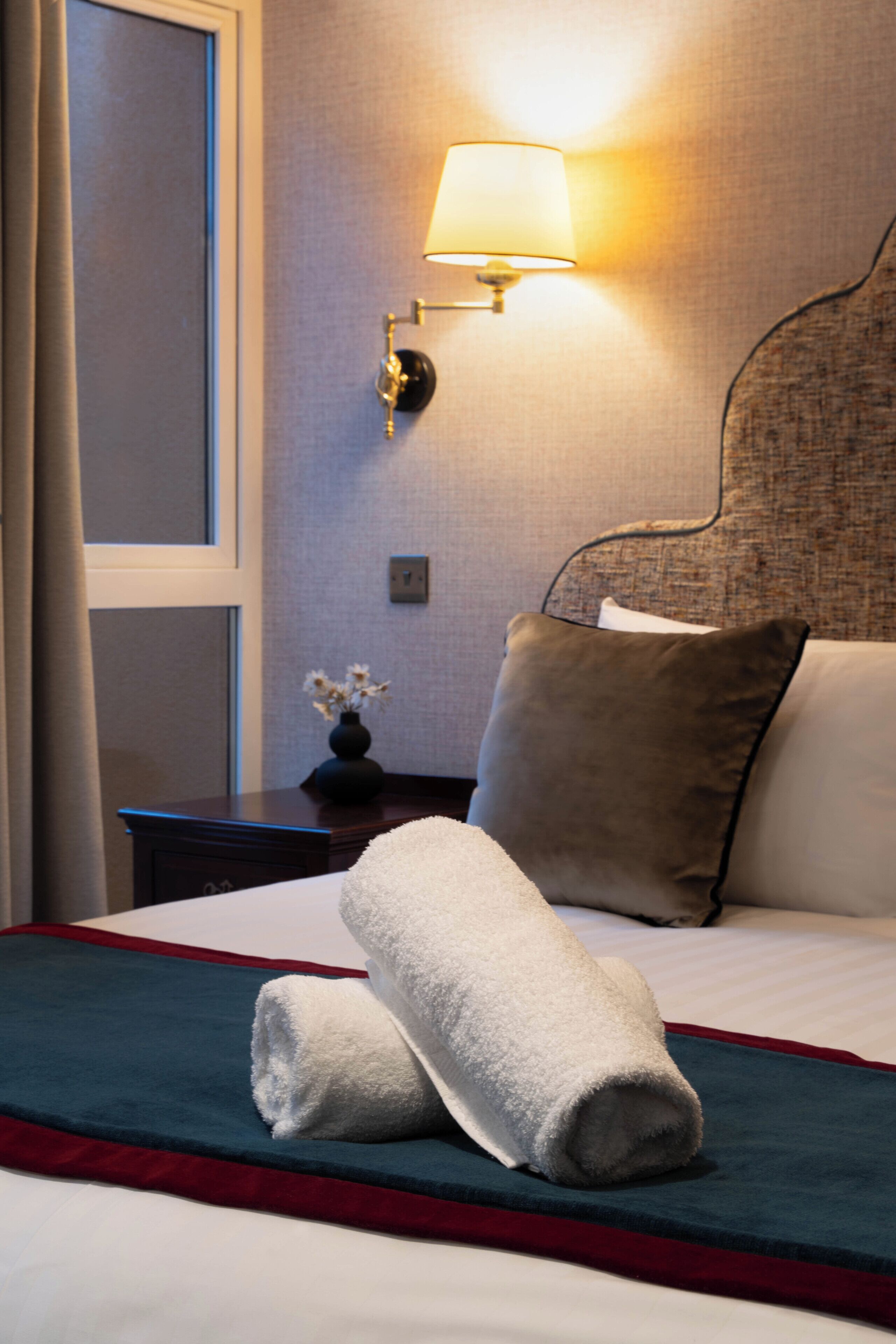 A neatly made bed with a white and blue bedspread, a pair of rolled towels, and a nightstand with a lamp.