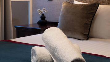 A neatly made bed with a white and blue bedspread, a pair of rolled towels, and a nightstand with a lamp.