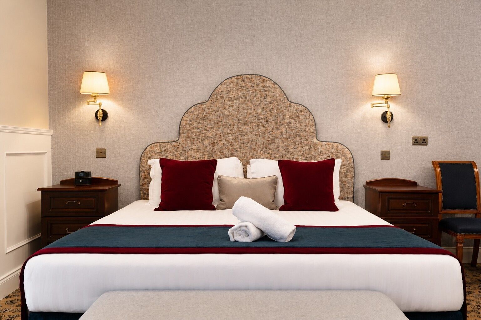 Quarto de hotel com uma cama grande, duas mesas de cabeceira, cabeceira com padrão texturizado e luminárias fixadas na parede.