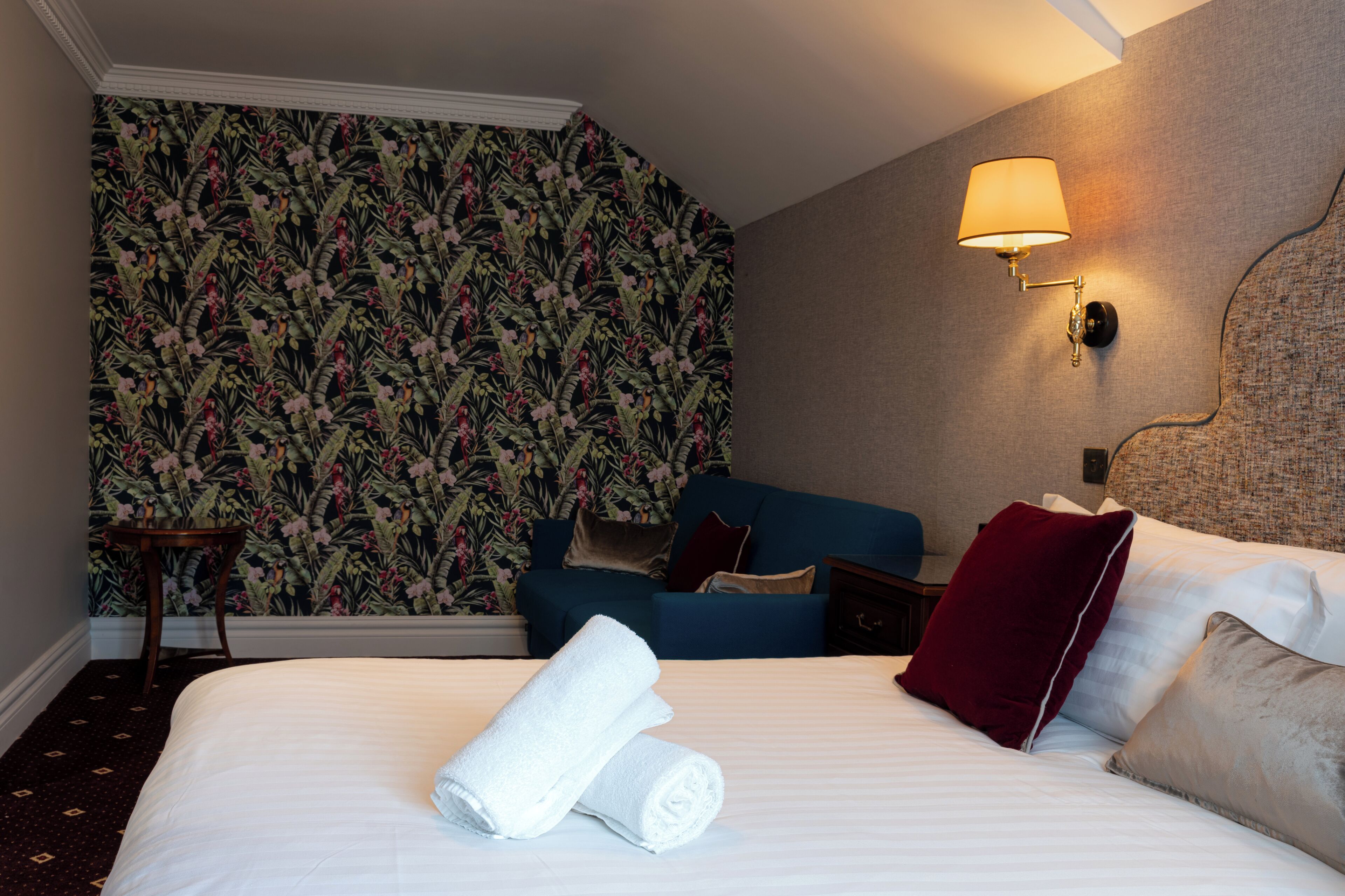 A hotel room with a bed, a blue sofa, a nightstand, a lamp, and a floral wallpaper.