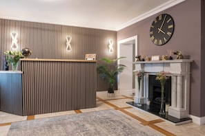 A reception area with a wooden reception desk, a large wall clock, and a fireplace with decorative items. - THE VÉRO (Killarney)