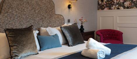 A hotel room with a large bed, multiple pillows, a red armchair, a wall-mounted lamp, and a floral wallpaper.