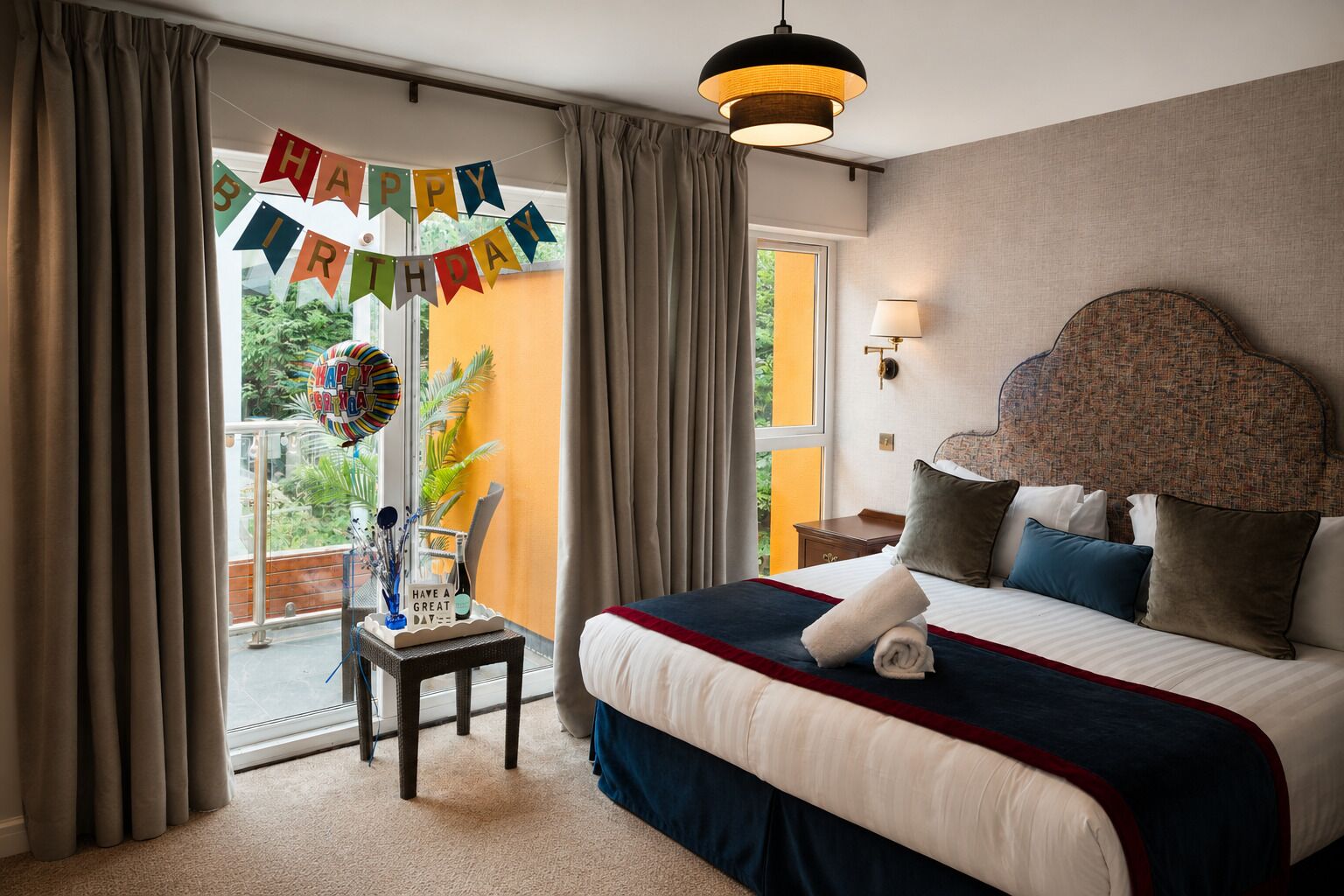 A hotel room with a bed, a nightstand, a lamp, a window with curtains, and a balcony with a birthday banner.
