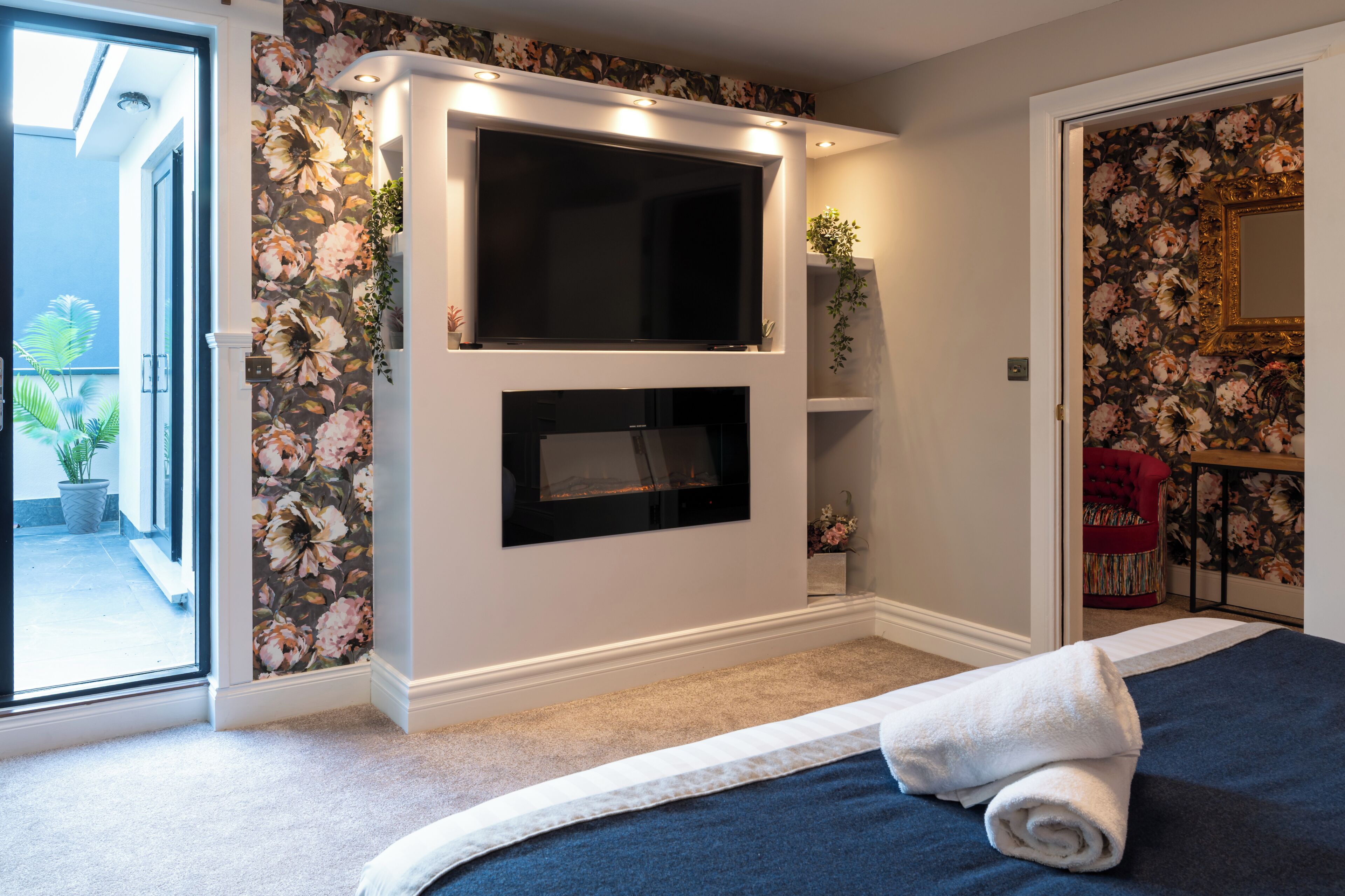 A bedroom with a large bed, a flat-screen TV mounted on the wall, and a fireplace below the TV.