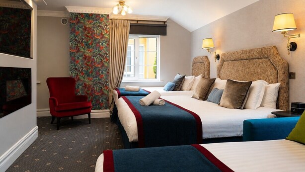 Une chambre d’hôtel avec deux lits, un fauteuil rouge, une grande fenêtre avec des rideaux et des lampes fixées au mur.