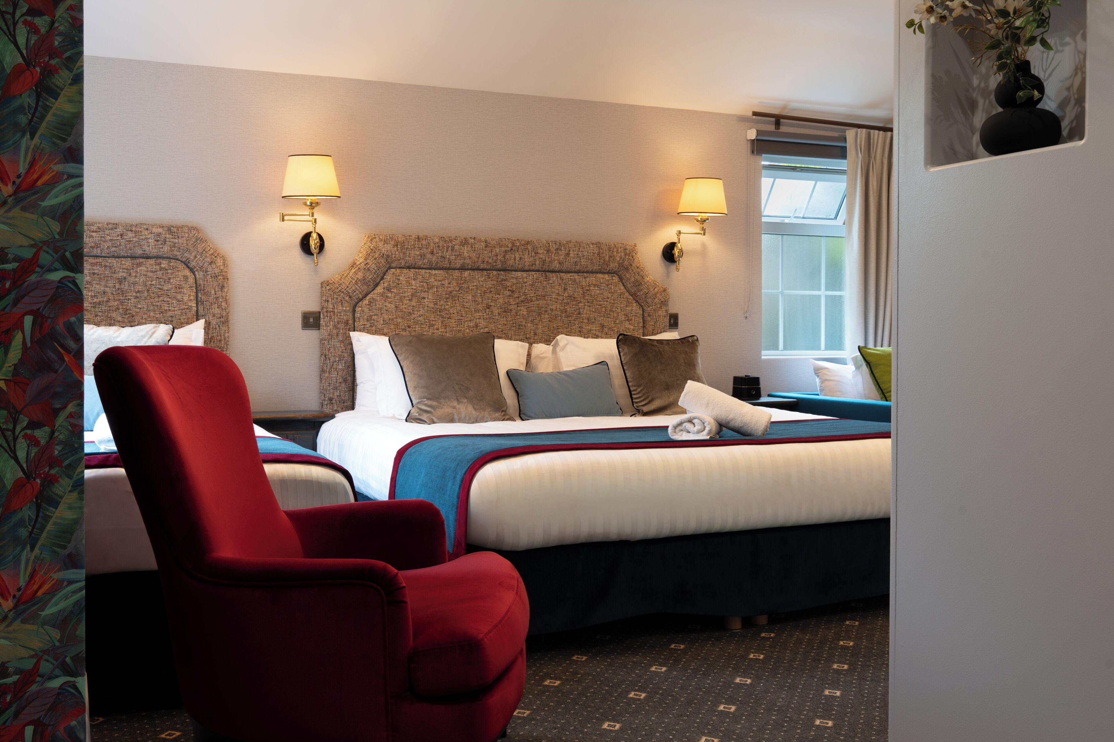 Habitación de hotel con cama, un sillón rojo, dos lámparas de pared y un ventanal con cortinas.