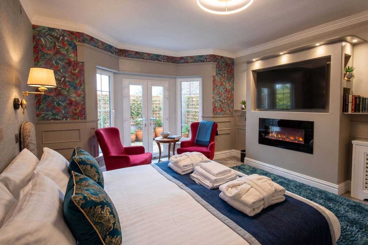A hotel room with a bed, two red chairs, a fireplace, a TV, and a view of the outside.
