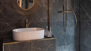 A modern bathroom with a marble sink, a sunburst mirror, a walk-in shower, and a wooden shelf with towels.