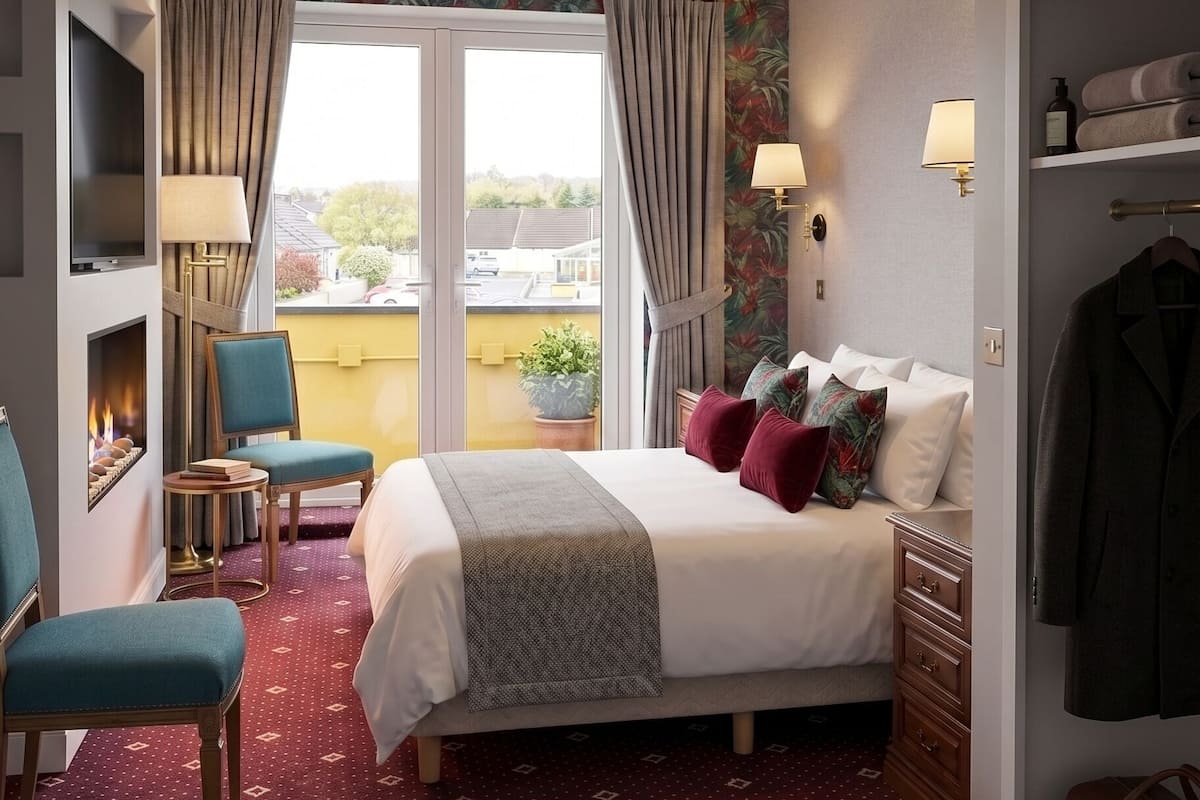 A hotel room with a bed, a seating area with chairs, a TV, a fireplace, a balcony with curtains, and a wardrobe.
