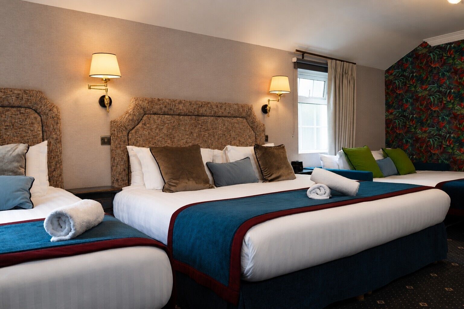 Una habitación de hotel con dos camas, cada una con una colcha azul y roja, ropa de cama blanca y varias almohadas. Hay dos lámparas de pared a cada lado de las camas.