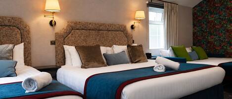 Una habitación de hotel con dos camas, cada una con una colcha azul y roja, ropa de cama blanca y varias almohadas. Hay dos lámparas de pared a cada lado de las camas.