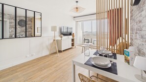 A modern living room with a wooden floor, a dining table set with plates and glasses, a TV, and a large window with a view. - Sea view for 4 by the beach (Saint-Hilaire-de-Riez)