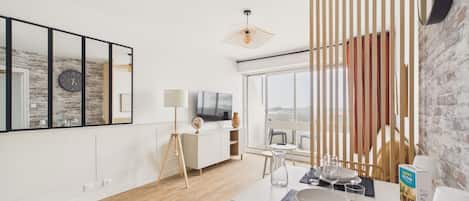 A modern living room with a wooden floor, a dining table set with plates and glasses, a TV, and a large window with a view.