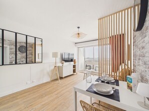 A modern living room with a wooden floor, a dining table set with plates and glasses, a TV, and a large window with a view. - Sea view for 4 by the beach (Saint-Hilaire-de-Riez)