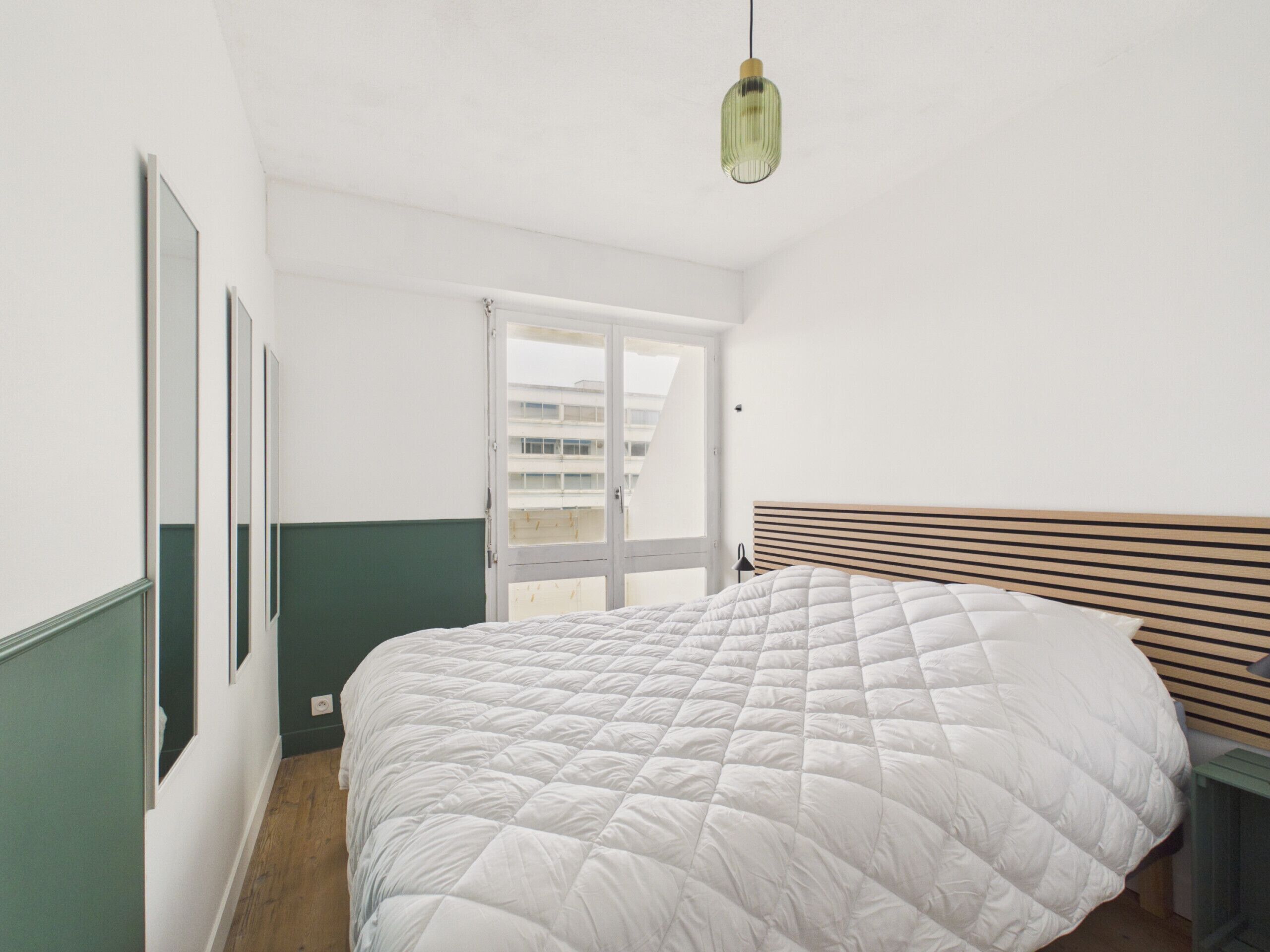Un dormitorio con cama, cabecera de madera, lámpara colgante y ventana con vistas a edificios.