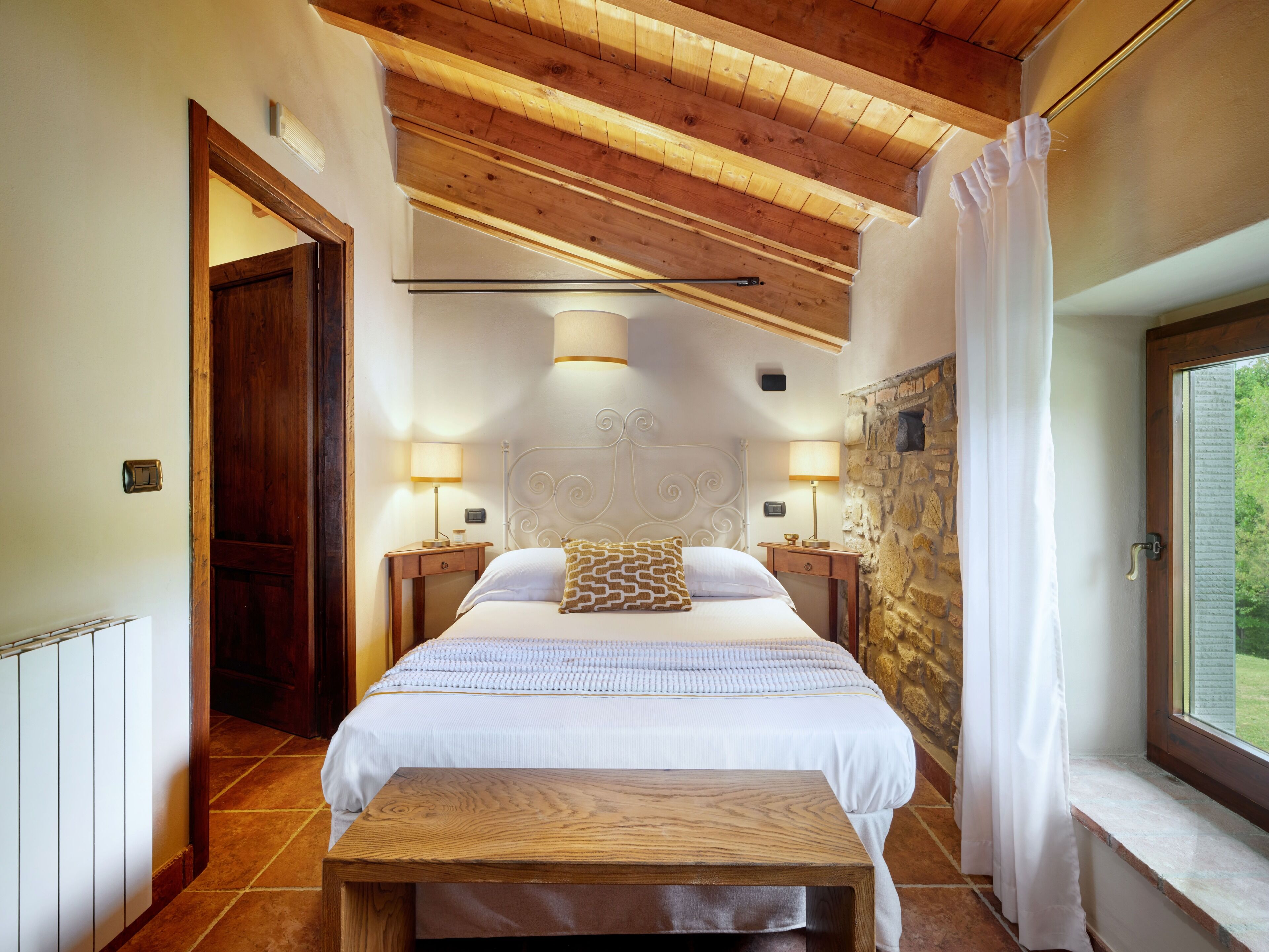 A bedroom with a wooden ceiling, a stone wall, a bed with white bedding, two bedside lamps, and a window with curtains.