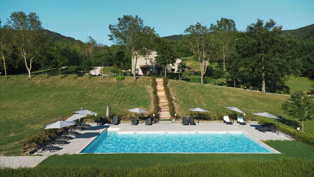 A pool with lounge chairs and umbrellas, surrounded by a grassy area and trees, leading to a stone building. - Tenuta Valdea (Firenzuola)