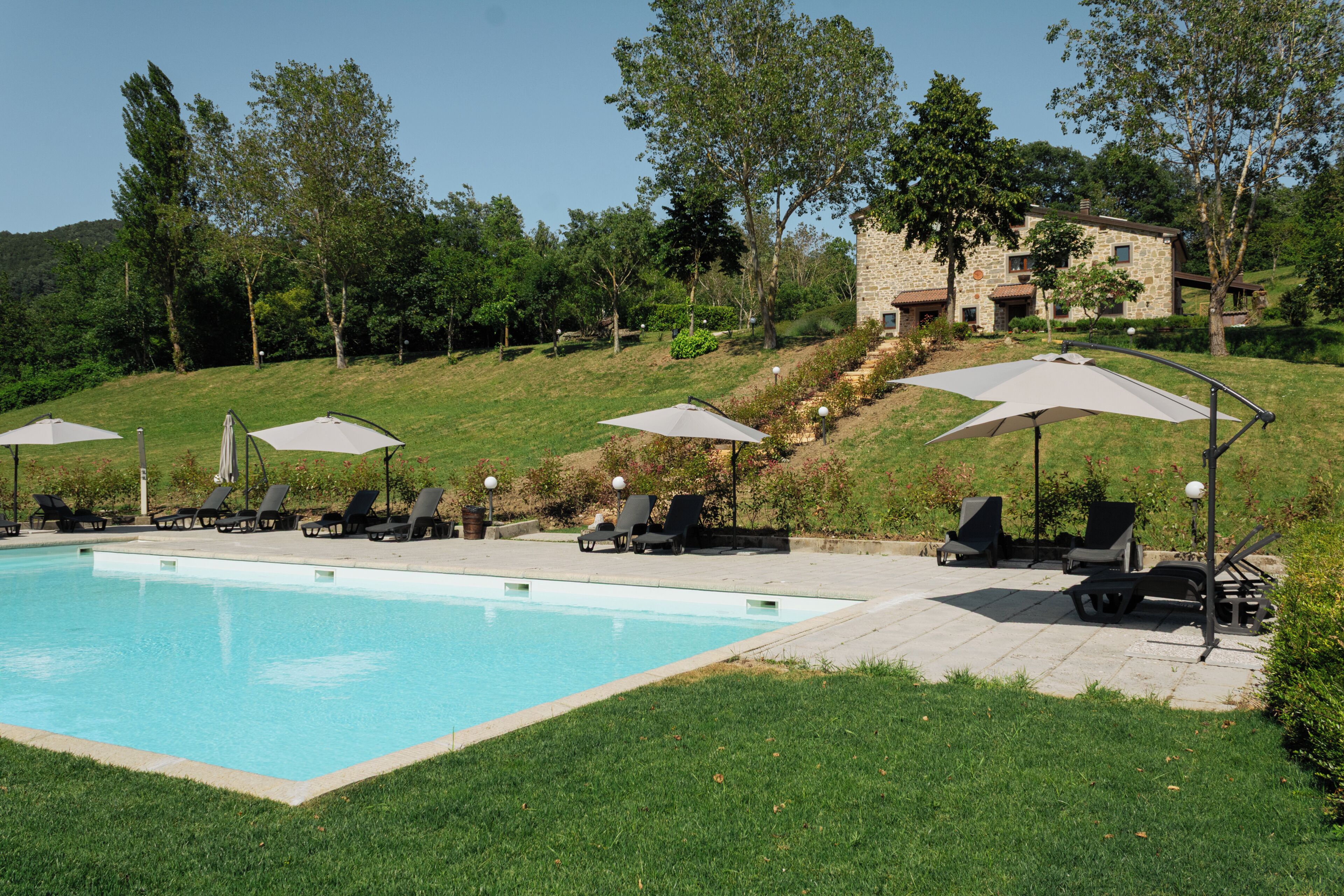 Piscina com espreguiçadeiras e guarda-sóis, cercada por vegetação e um edifício de pedra.