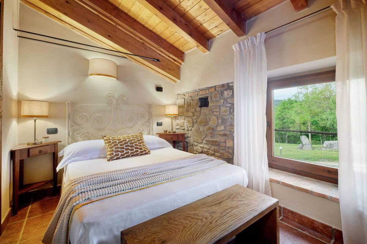 Un dormitorio con pared de piedra, vigas de madera en el techo, cama con almohada estampada, lámparas de noche y ventana con vistas al follaje.