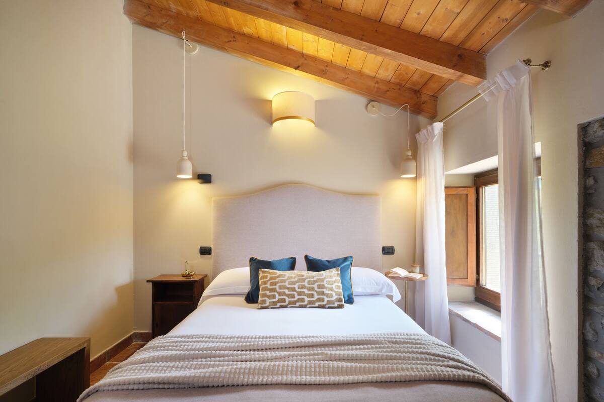 Un dormitorio ordenado con cama, lámparas de noche, mesita de noche y ventana con cortinas.
