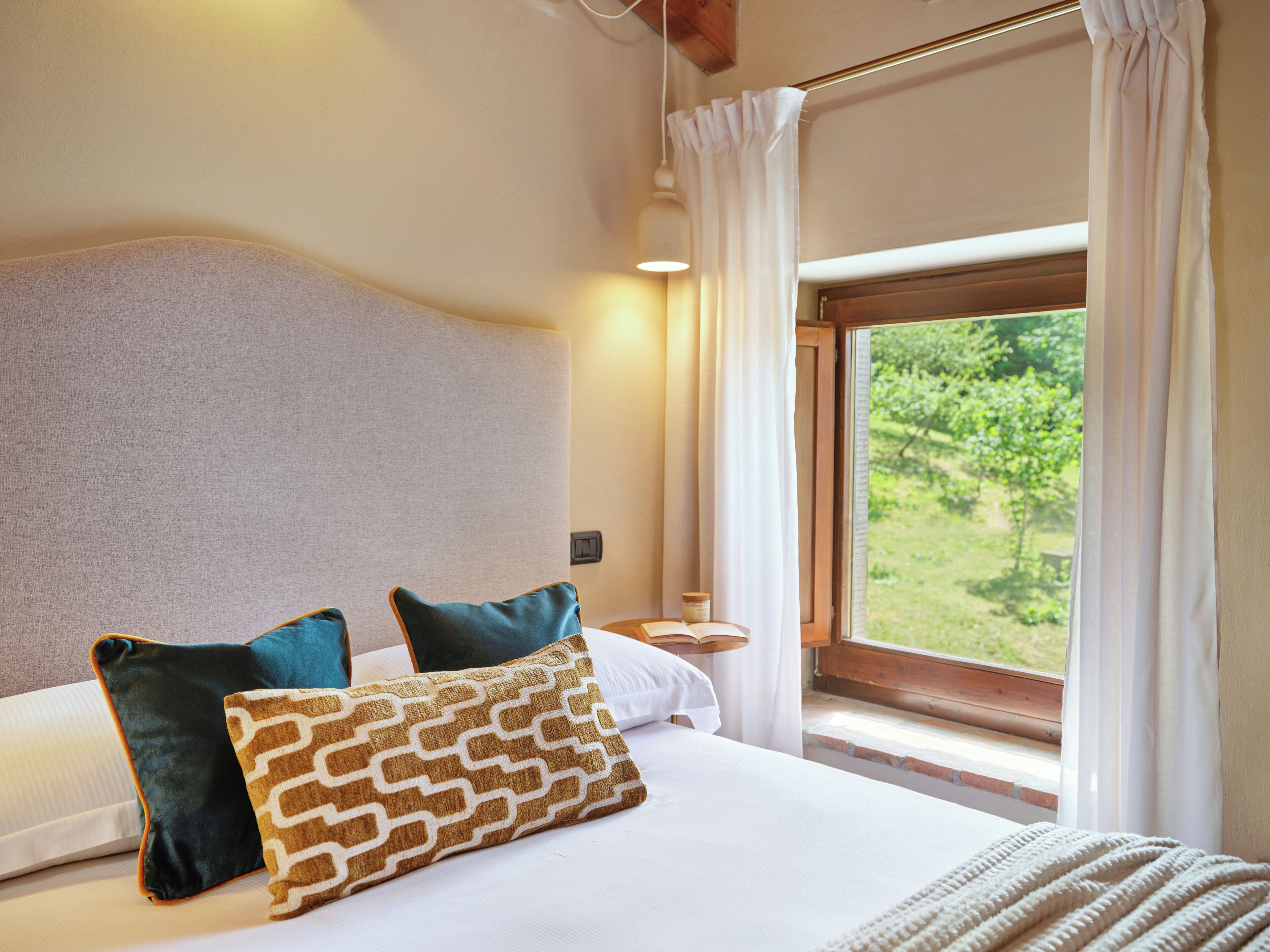 A bedroom with a bed, pillows, a bedside table, a lamp, and a window with a view of greenery.