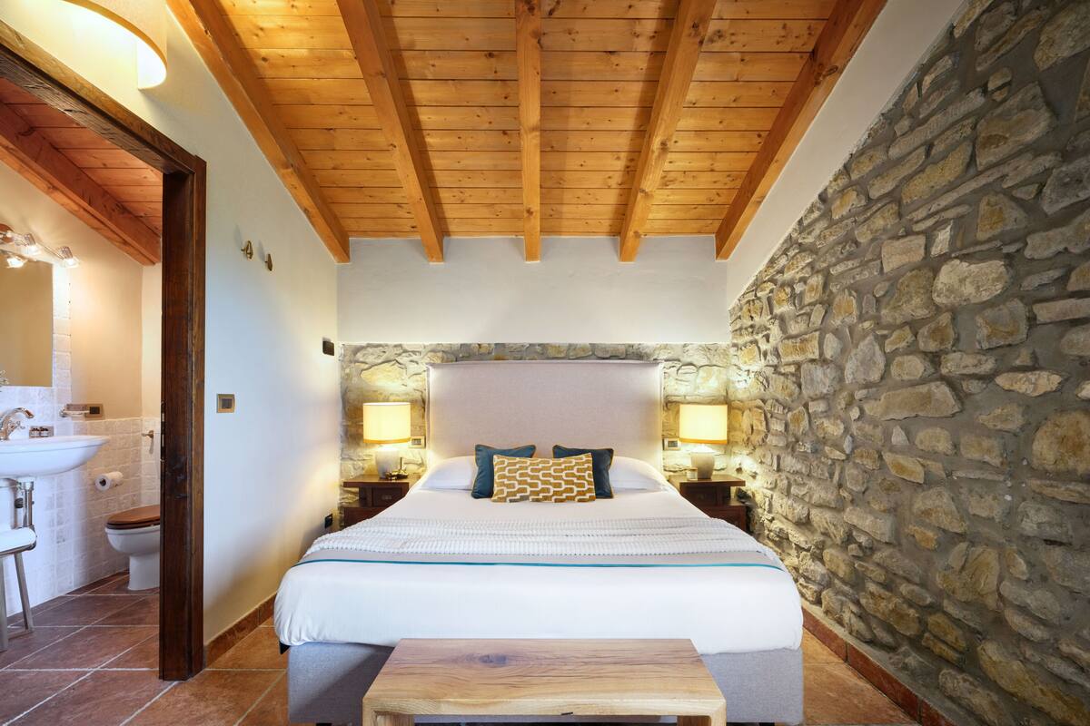 Un dormitorio con pared de piedra, vigas de techo de madera, una cama con almohadas, una mesita de noche y un taburete de madera.