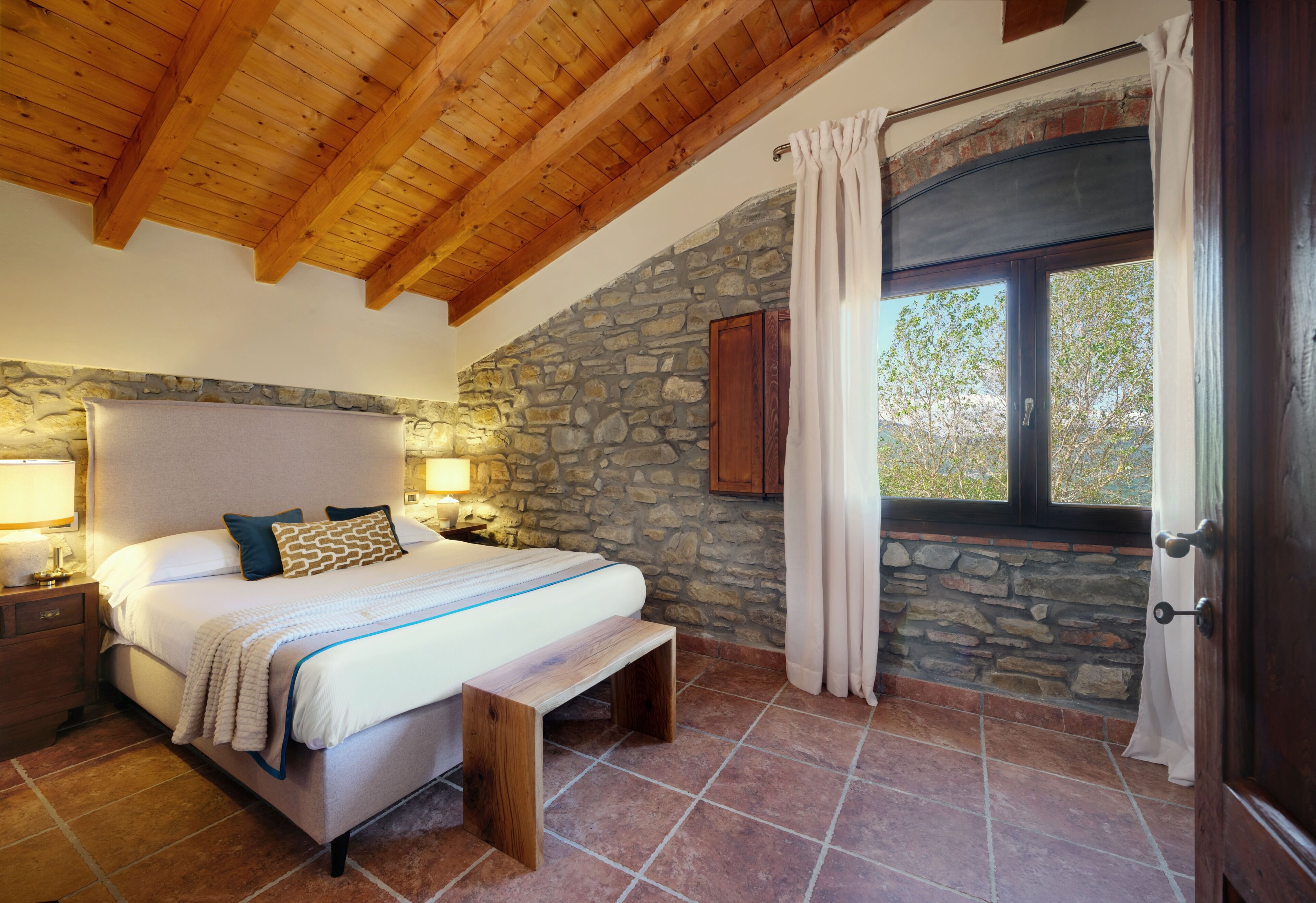 A bedroom with a stone wall, wooden ceiling beams, a bed with pillows, a bench, and a window with curtains.