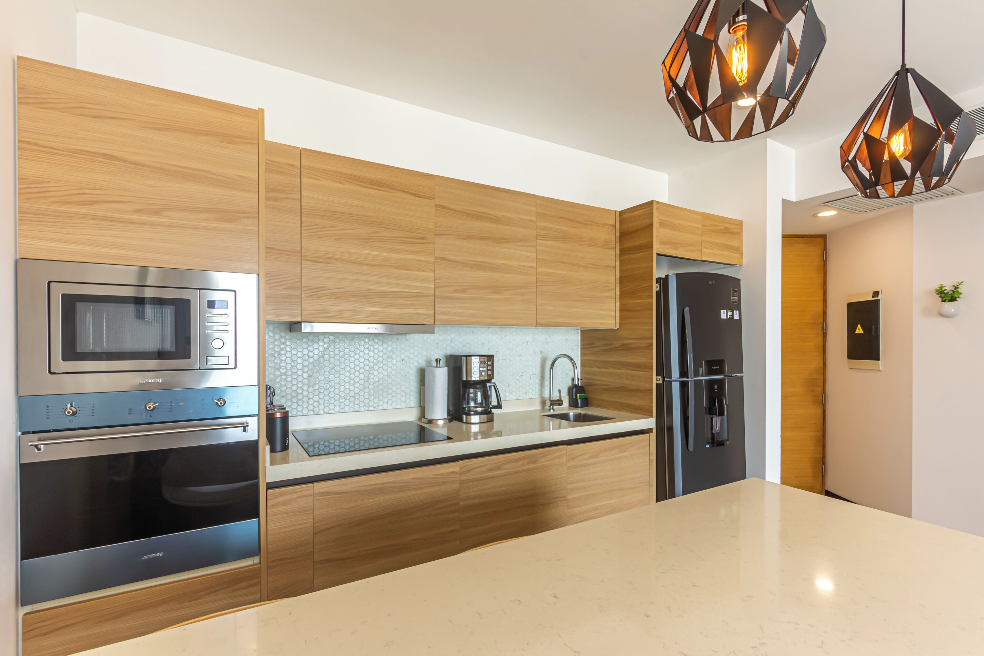 A modern kitchen with wooden cabinets, stainless steel appliances, a marble countertop, and geometric pendant lights.
