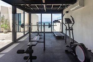 Ein Fitnessraum mit Trainingsgeräten, einer Bank und Meerblick durch große Fenster.