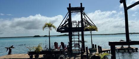 A wooden dock extending into calm water, a wooden structure with chairs, and a clear sky.