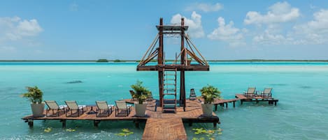 A wooden pier leading to a structure with a ladder, surrounded by lounge chairs and potted plants, overlooking clear turquoise waters.