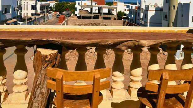 Un balcon avec des chaises en bois, une vue sur les immeubles et un ciel dégagé.