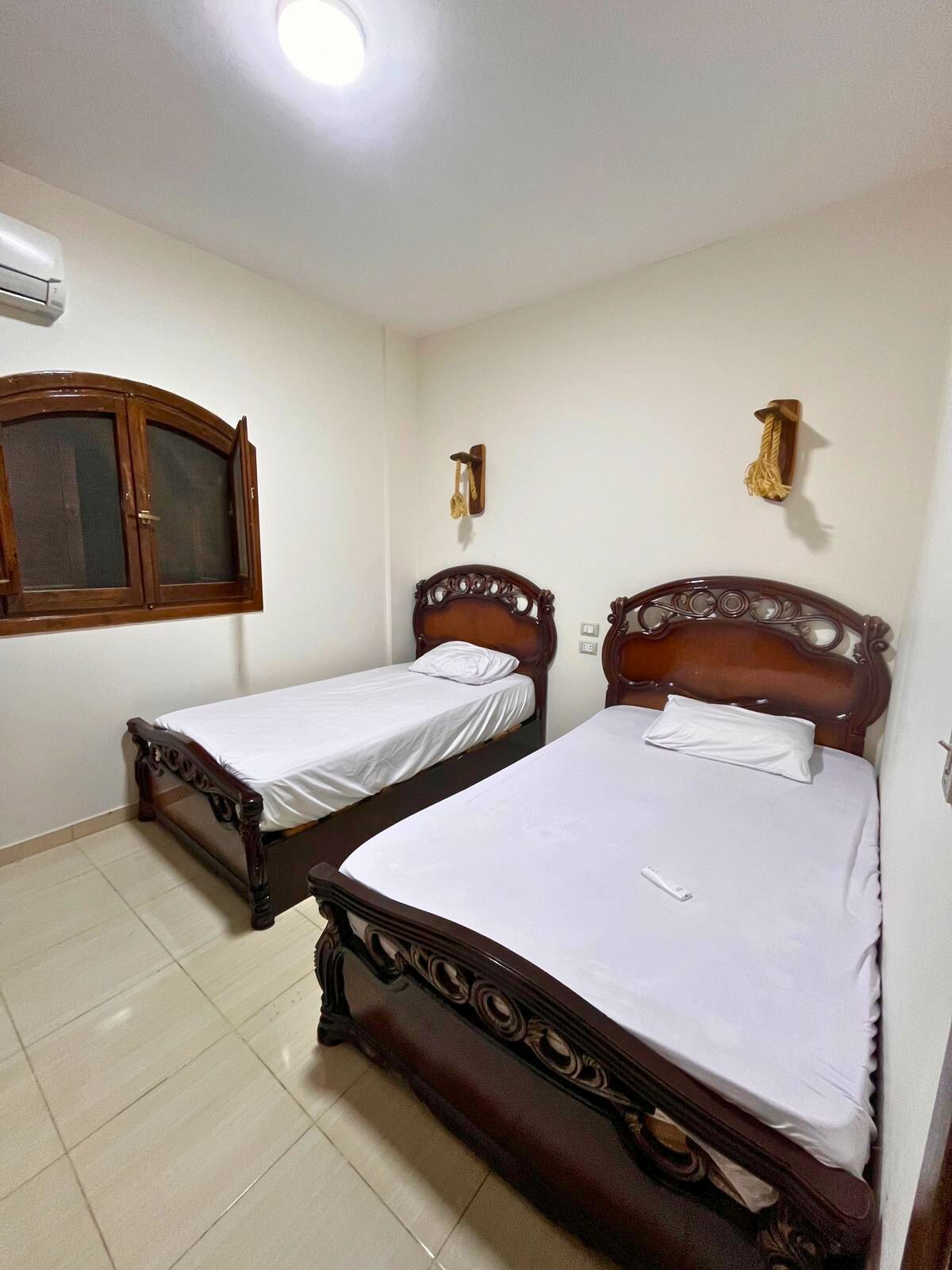 A room with two wooden beds, a window, a wall-mounted air conditioner, and a light fixture.