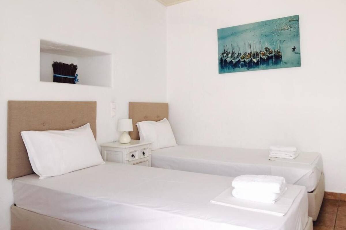 A hotel room with two beds, a painting of boats, a small shelf, and a nightstand with a lamp.