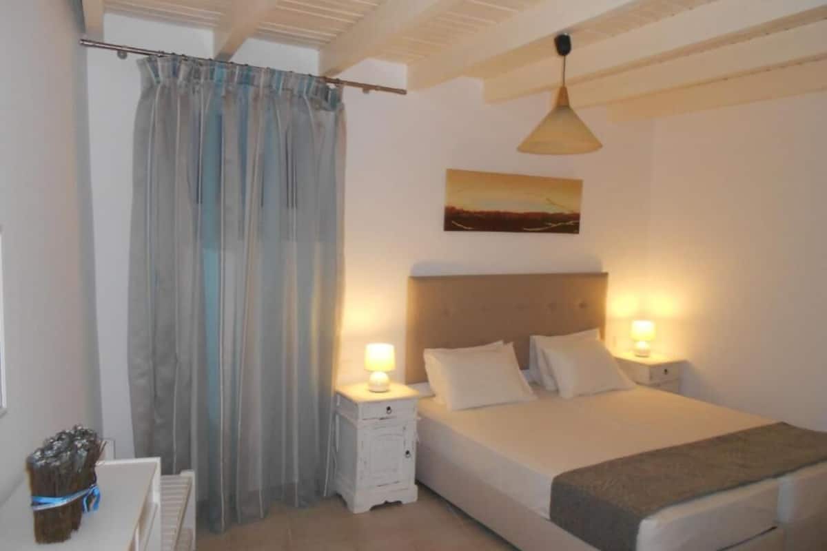 A bedroom with a bed, two bedside lamps, a nightstand, a window with curtains, and a painting on the wall.