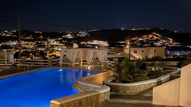 A swimming pool with a curved edge, surrounded by stone walls and a small garden area with palm plants. - Casa Bianca Mykonos (Ornos)